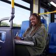 A student sits in a bus seat, looking down at the books on their lap and smiling.