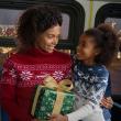 A child sits on a adult's lap inside a bus. They are dressed festively and carrying a wrapped gift. The neighborhood seen through the bus windows is decorated with lights.