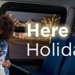 A child and an adult in festive sweaters smile at each other on a bus. The adult has a wrapped gifts. Trees seen through the bus windows are decorated with lights. Text says: Here for the holidays