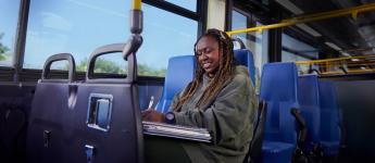 A student sits in a bus seat, looking down at the books on their lap and smiling.
