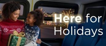 A child and an adult in festive sweaters smile at each other on a bus. The adult has a wrapped gifts. Trees seen through the bus windows are decorated with lights. Text says: Here for the holidays