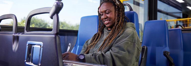 A student sits in a bus seat, looking down at the books on their lap and smiling.