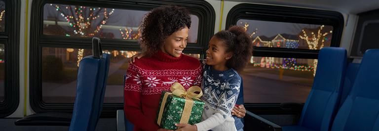 A child sits on a adult's lap inside a bus. They are dressed festively and carrying a wrapped gift. The neighborhood seen through the bus windows is decorated with lights.