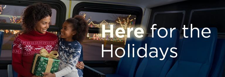 A child and an adult in festive sweaters smile at each other on a bus. The adult has a wrapped gifts. Trees seen through the bus windows are decorated with lights. Text says: Here for the holidays